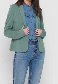 Grön skräddarsydd kavaj med slagkrage, lätt tyg och enkelknappsstängning, bärs över en ljus denimskjorta och högmidjade jeans.