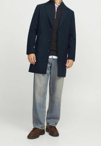 Herrjacka i mörkgrönt med skräddarsydd design, svart tröja under, ljusblå skjorta, ljusa denimjeans och bruna skor med tjock sula.