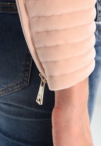 Jasnoróżowa kurtka zapinana na zamek z prążkowaną teksturą na rękawie, z błyszczącym złotym zamkiem i deninowym tłem.