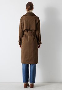 Bruine trenchcoat met een ceintuurgordel in de taille en schouderepauletten, gecombineerd met blauwe jeans en beige blockhakken.