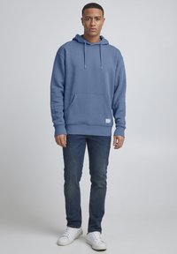 Felpa blu con tasca frontale e cappuccio con cordoncino, realizzata in tessuto liscio. Abbinata a jeans scuri e sneakers bianche.