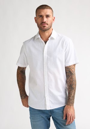 Mann mit Tätowierungen trägt ein weißes kurzärmeliges Hemd mit Knöpfen und blaue Jeans, steht vor einem einfachen hellen Hintergrund.