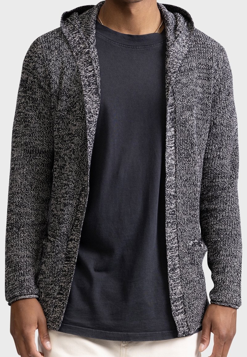 Homme portant un t-shirt noir sous un cardigan à capuche gris et noir, tricoté, ouvert à l'avant avec des poches.