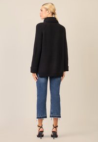 Maglione oversized nero a coste con colletto alto, abbinato a jeans blu cropped con orli sfilacciati e décolleté neri con cinturini.