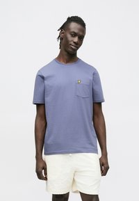 Maglietta di cotone viola con scollo rotondo, caratterizzata da un piccolo logo giallo sulla tasca del petto; abbinata a pantaloncini di colore crema con trama.