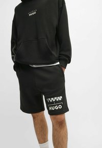 Sweat à capuche noir avec logo sur le devant et détail sur le côté, associé à un short noir à motif à carreaux et logo sur la cuisse.
