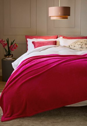 Lit avec une literie blanche et une couverture en velours rose vif, des coussins roses et à motifs, une lampe beige suspendue et des fleurs roses sur la table d'appoint.