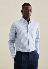 Camicia a maniche lunghe da uomo con strisce verticali blu e bianche, chiusura con bottoni, colletto classico e tessuto testurizzato, abbinata a pantaloni scuri.