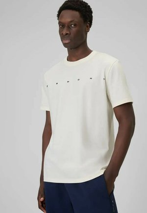 Man draagt een crème kortärmelig T-shirt met zwarte letters met ruimte ertussen en donkerblauwe broek, staand met één hand in de zak.