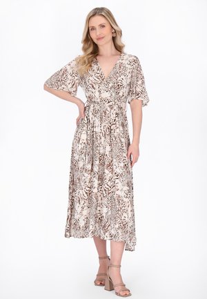Donna con capelli biondi ondulati che indossa un vestito midi beige e marrone con stampa animale, in piedi con una mano sui fianchi e sandali beige con tacco a blocco.