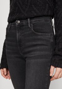 Jeans de mezclilla negra de tiro alto con diseño de cinco bolsillos, desvanecidos sutilmente y cierre de botón metálico. Combinados con un suéter de punto negro texturizado.