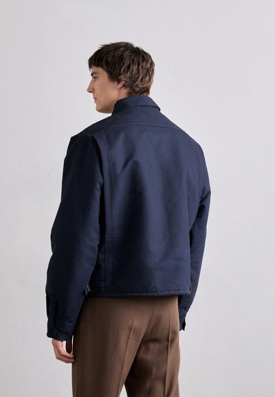 Navy blue jacket in thick fabric, featuring a high collar and side pockets, worn over brown trousers. Simple, clean design.
