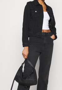 Persona vistiendo una chaqueta de mezclilla negra y jeans con una camiseta corta blanca, sosteniendo un bolso negro por la correa con una mano.