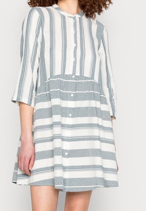 Woman wearing a knee-length dress with vertical and horizontal blue and white stripes and three-quarter sleeves, buttons down the front.