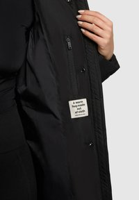 Interior de una chaqueta acolchada negra con un bolsillo con cremallera y una etiqueta de tela que dice: "Un abrazo cálido hecho de tela." Textura suave, sin patrones.