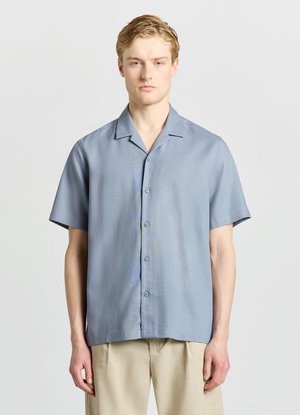 Jeune homme aux cheveux blonds courts porte une chemise bleu clair à manches courtes boutonnée et un pantalon beige sur un fond uni.