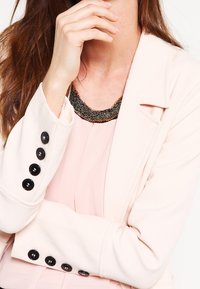 Blazer rosa claro con un diseño ajustado, botones negros y un cuello texturizado que presenta un acento oscuro con cuentas. Llevado sobre una blusa rosa.