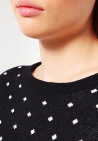 Czarny dziany sweter w białe kropki, z okrągłym dekoltem i prążkowanym kołnierzem. Miękka faktura widoczna wokół dekoltu.