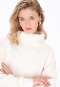 Cremefarbener Pullover mit hohem Rippenkragen, weicher Textur und langen Ärmeln. Das Modell hat die Arme verschränkt und zeigt eine lockere Passform.