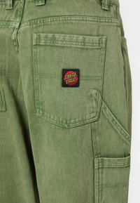 Groene katoenen cargobroek met meerdere zakken en een zwart logo-patch met "SANTA CRUZ" in geel en rood. Het gemêleerde materiaal is zichtbaar.