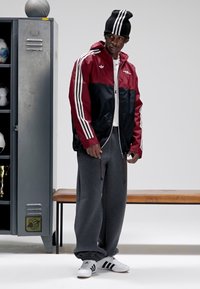 Rød og svart jakke med glidelås og hvite striper, grå joggebukser og hvite joggesko. Svart lue fullfører det atletiske utseendet.