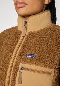 Gilet en polaire marron avec col haut, fermeture éclair à l'avant et poche sur la poitrine. Présente le logo Patagonia. Matériau extérieur texturé et fermetures éclair lisses.