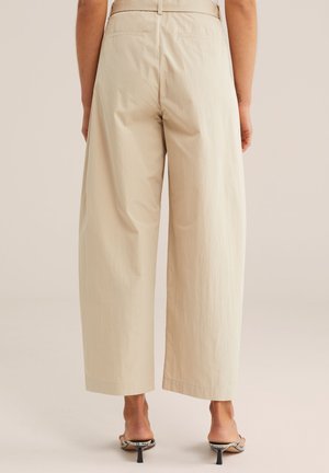 Beżowe spodnie o wysokim stanie i szerokich nogawkach noszone przez osobę stojącą, zestawione z czarnymi sandałami na obcasie z paskami w wężowy wzór.