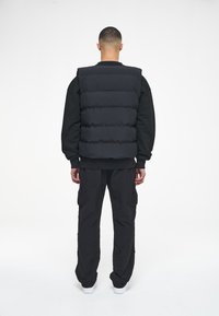 Svart pufferväst med horisontell sömnad och hög krage, bärs över en svart sweatshirt och kombineras med löst sittande svarta cargo-byxor.