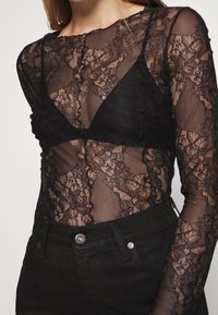 Czarna koronkowa bluzka z długim rękawem i kwiatowymi wzorami, noszona na ciemnym biustonoszu. Posiada przezroczystą tkaninę i okrągły dekolt. Połączona z czarnymi dżinsami.