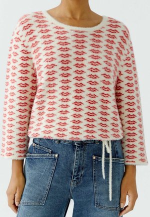 Osoba ubrana w puszysty biały sweter z czerwonymi wzorami w kształcie ust oraz niebieskie dżinsy z dużymi przednimi kieszeniami i sznurkiem do wiązania w talii.