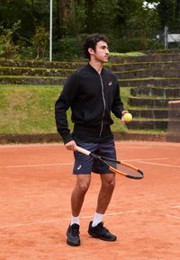 Mann in schwarzer Jacke und blauen Shorts, der einen Tennisschläger und einen Ball auf einem Platz mit Tonboden hält, wobei Steinstufen und Grün im Hintergrund zu sehen sind.