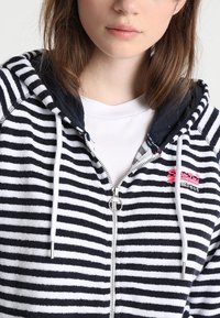 Ritsvest met marineblauwe en witte strepen, gemaakt van zachte badstof, met een marineblauwe capuchon, witte trekkoorden en een roze logo op de borst.
