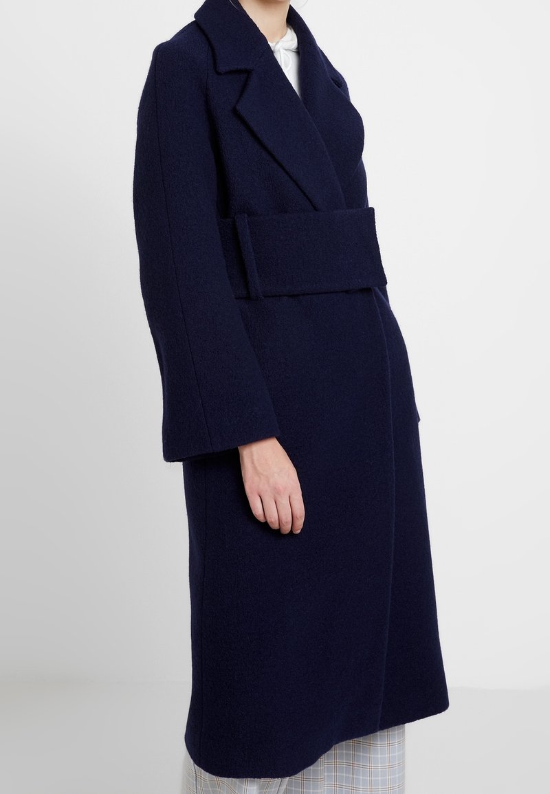Marineblauer Wollmantel mit Wickeldesign, breiten Revers, langen Ärmeln und einem passenden Gürtel aus dem gleichen Stoff. Die Textur ist weich und leicht flauschig.
