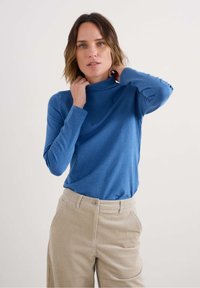 Mėlyna ilgomis rankovėmis turtlenecks marška, pagaminta iš minkštos medžiagos, derinama su smėlio spalvos kordo kelnių su vertikaliu braižymu ir priekinėmis kišenėmis.