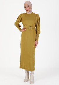 Vestido de punto de manga larga en verde mostaza, con cintura ceñida por un cinturón y mangas abullonadas. Combinado con botas blancas de tacón. Textura suave.
