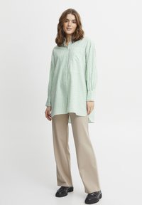 Camicia a maniche lunghe a righe verdi e bianche con taschino, abbinata a pantaloni beige a gamba larga e scarpe nere, indossata da un modello su uno sfondo bianco.