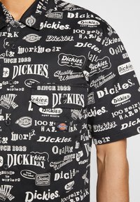 Chemise noire à manches courtes recouverte de divers logos et textes blancs Dickies, avec un bouton sur le devant et une poche poitrine.