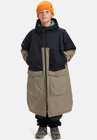 Jong persoon in een lange oversized zwart en beige jas, oranje beanie, zwarte broek en grijze sneakers, staande met handen in de zakken.