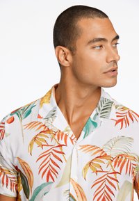 Camisa de manga corta con una base blanca y patrones de hojas tropicales coloridas en rojo, naranja, amarillo y verde; diseño de botones en el frente.