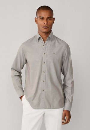 Camisa de manga larga gris claro con textura sutil, que presenta un pequeño logo bordado y botones marrones. Combinada con pantalones blancos.