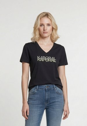 Blonde vrouw met een zwart V-hals T-shirt met de tekst "KAPORAL" en blauwe spijkerbroek, staand tegen een effen lichte achtergrond.