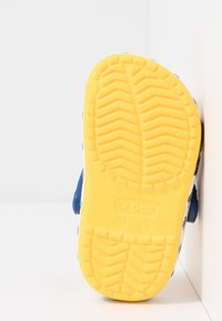 Blå sko med en texturerad gul gummisula, prydd med ett zickzackmönster och varumärkeslogotyp. Vit ovandel med färgglada blommönster.