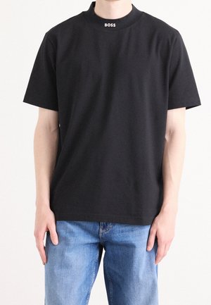 Homme portant un T-shirt noir à manches courtes avec le texte "BOSS" sur le col et un jean bleu, debout devant un fond blanc.