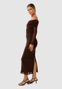 Robe marron à épaules dénudées avec détails froncés, silhouette ajustée et fente latérale. Associée à des talons noirs à lanières. Tissu lisse et extensible.