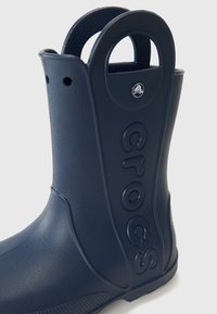 Stivale in gomma impermeabile di colore blu navy con una superficie testurizzata, ampie maniglie a forma di cerchio, e logo "crocs" impresso sul lato.