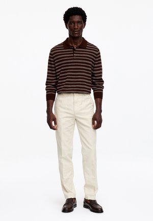 Homme debout portant un polo à manches longues rayé marron et gris, un pantalon crème et des mocassins marron foncé sur fond blanc.