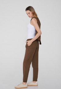 Braune Jogginghosen mit elastischen Bündchen und seitlichen Taschen, kombiniert mit einem weißen Tanktop und einem braunen Pullover, der über die Schulter hängt. Beigefarbene Turnschuhe.