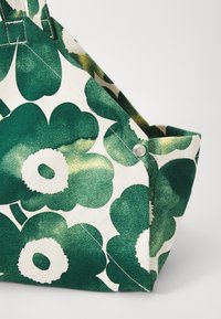 Canvas-Umhängetasche mit grünem Blumendesign, großen Blüten und hellen Akzenten. Mit einem silbernen Knopfdetail an der Seite.