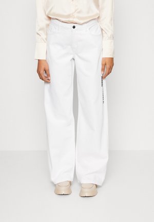 Persona con pantalones blancos holgados con texto en la pierna derecha, zapatos beige y una camisa de manga larga color crema, de pie frente a un fondo liso.