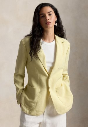 Mujer de cabello largo y oscuro, vestida con un blazer amarillo claro sobre una camisa blanca y pantalones blancos, de pie con las manos en los bolsillos.
